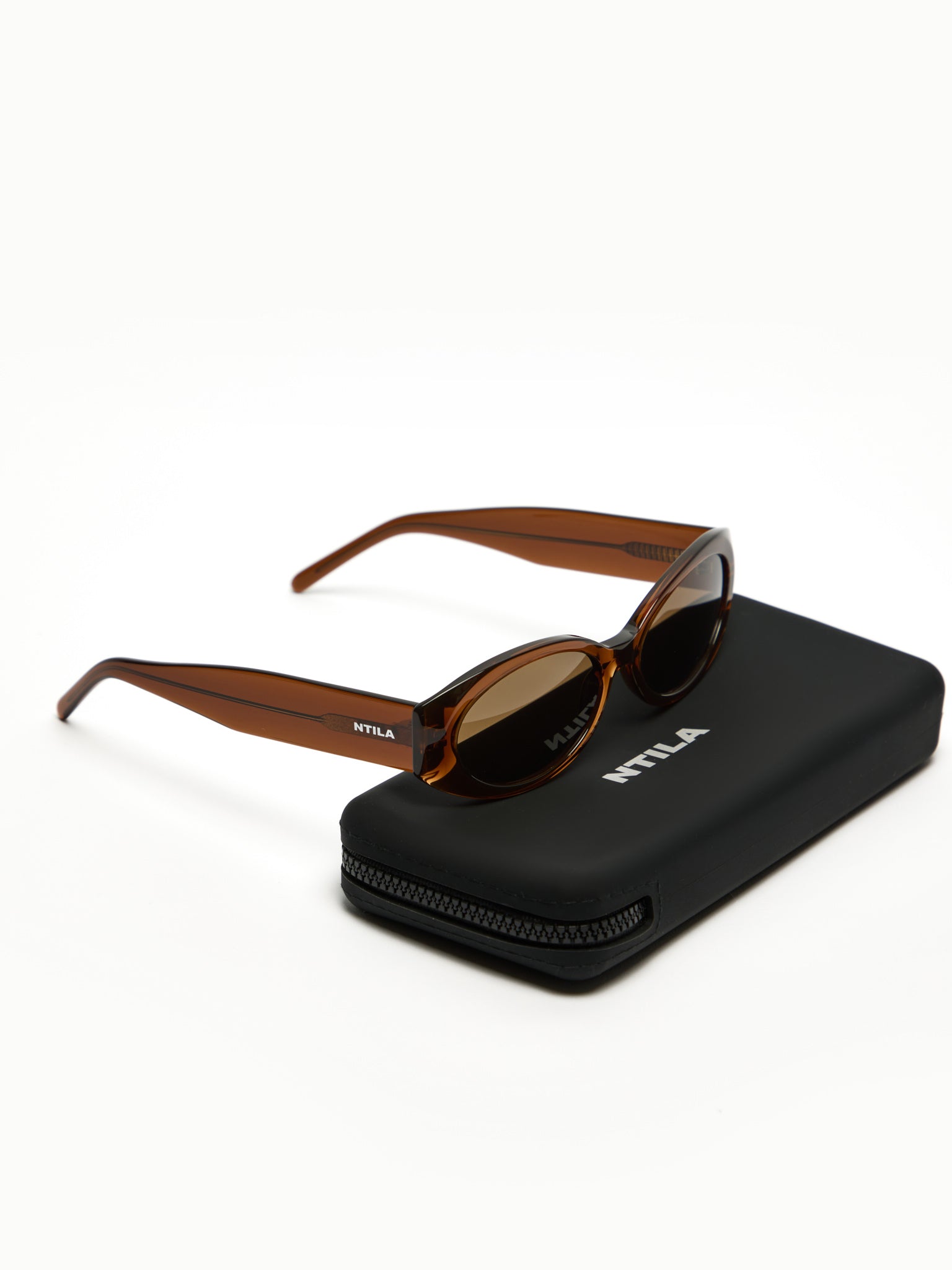 Cleo brown oval sunglasses with brown frame and dark lenses on a black case with 'NTILA' branding, on a white background.