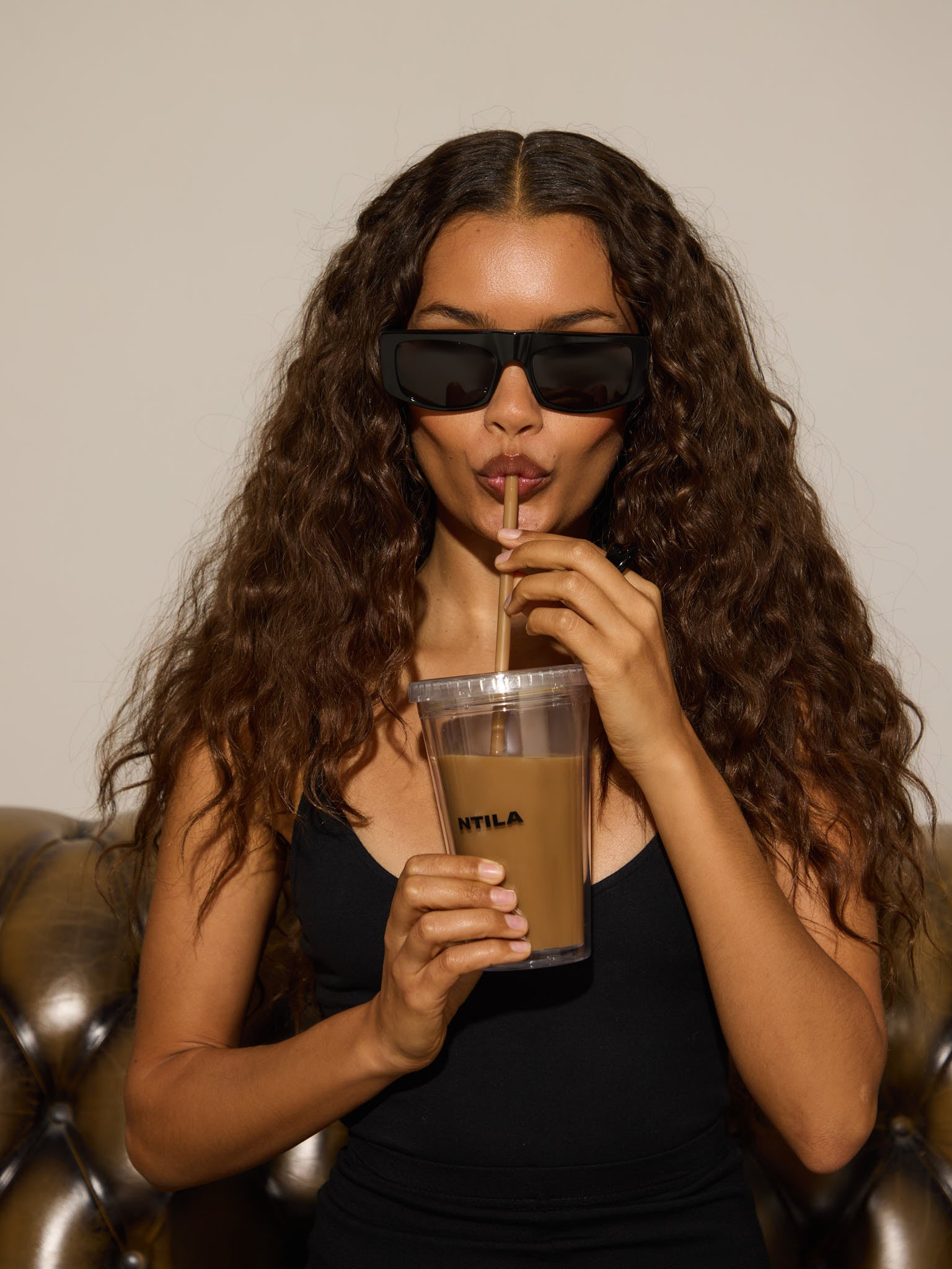 Woman drinking from a clear cup with a straw, wearing black oversized ntila sunglasses and a black top.